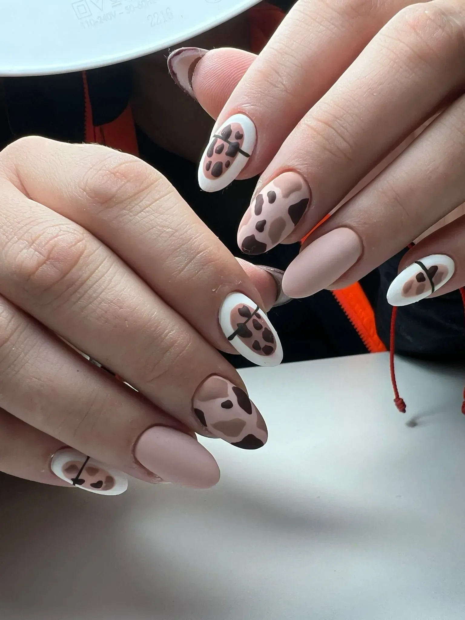 Manos con uñas ovaladas en color nude mate, algunas con diseño de estampado de vaca sobre puntas blancas.