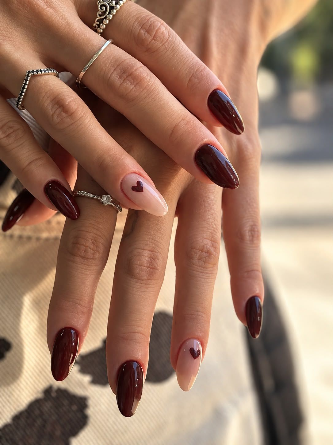 Manos con uñas largas color burdeos y nude, con una manicura cuidada y anillos delicados, posando al aire libre.