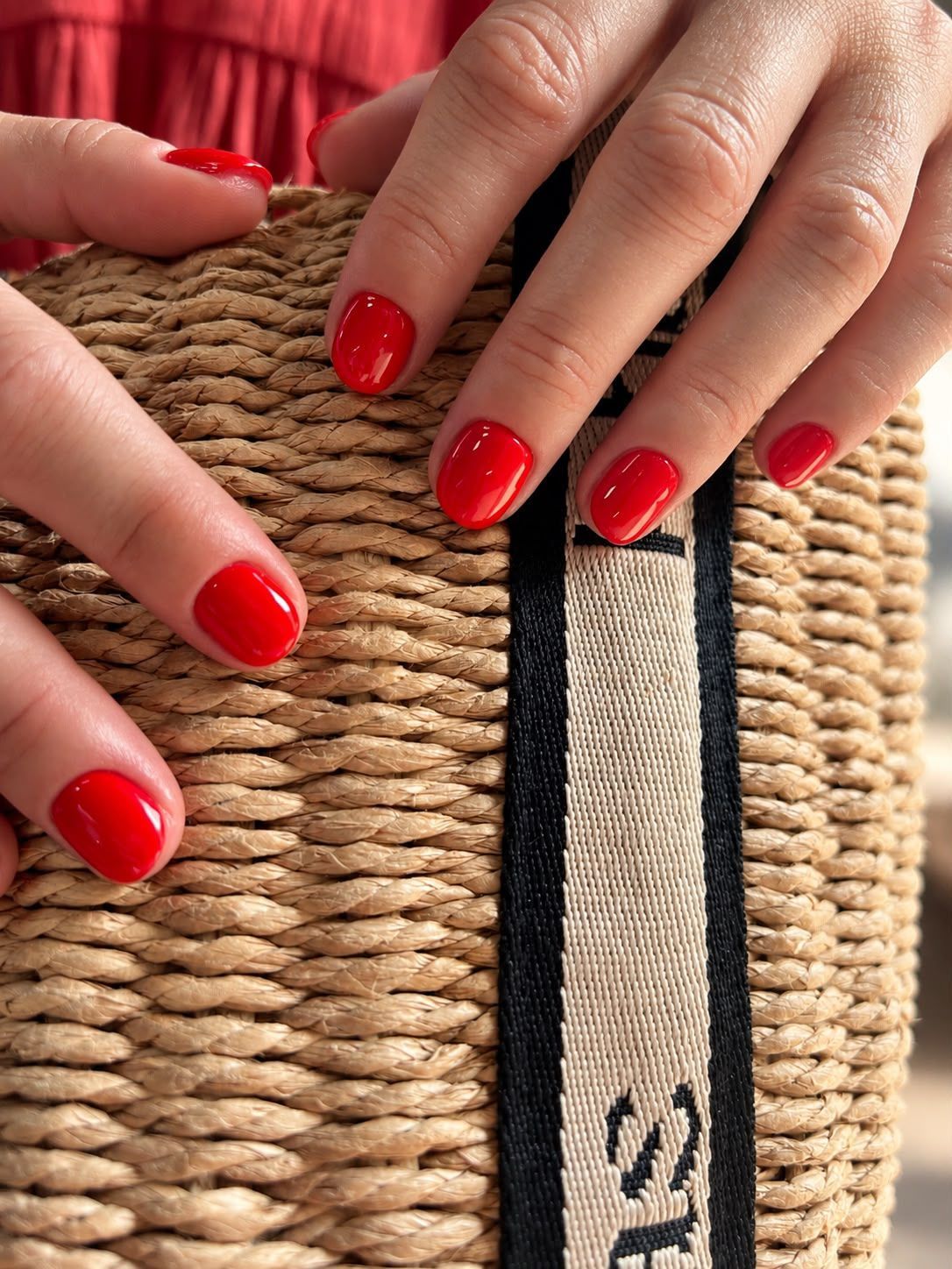 Unas manos con las uñas pintadas de rojo sostienen un bolso de paja trenzada con una correa en blanco y negro.