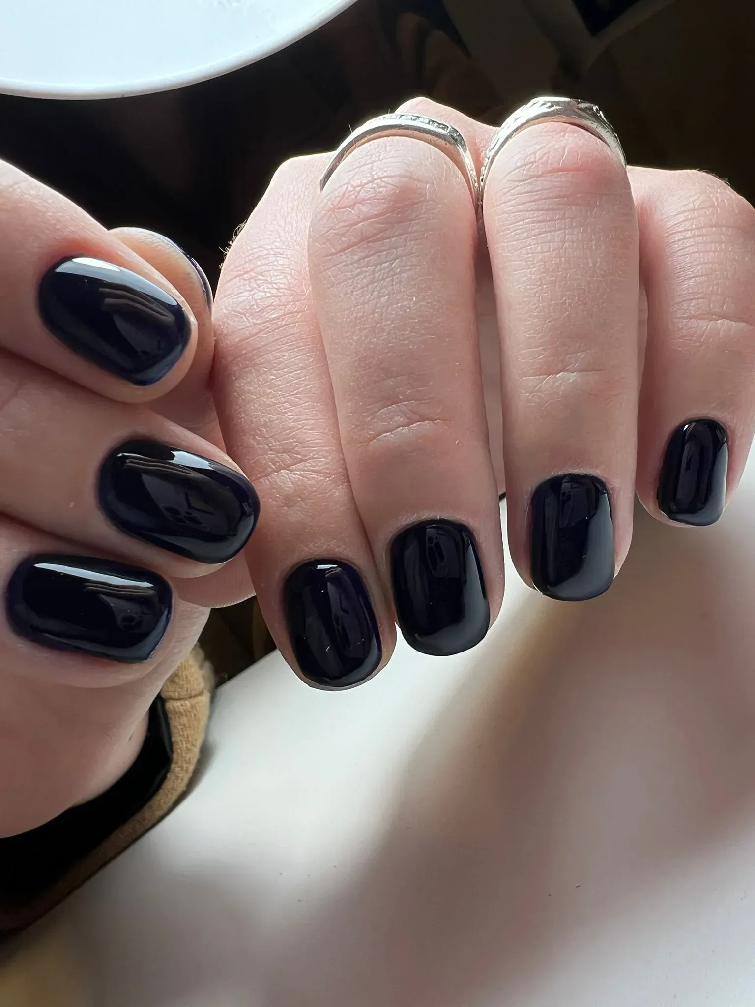 Mano con uñas ovaladas pintadas con esmalte negro brillante. Anillos de plata en dos dedos.