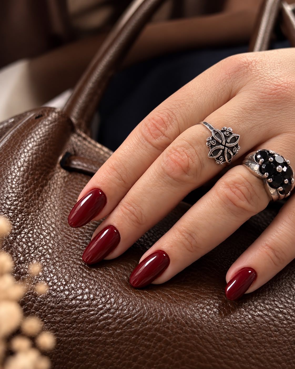 Mano con las uñas color burdeos apoyada sobre un bolso de cuero marrón, con anillos plateados y negros