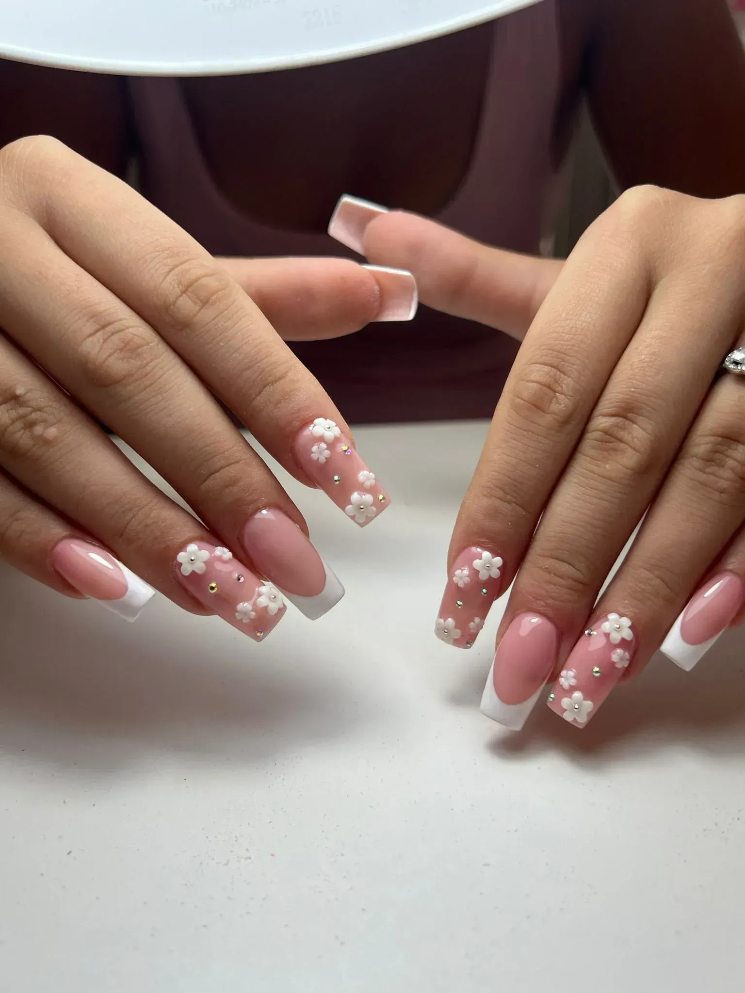 Manos con uñas acrílicas con punta francesa, algunas adornadas con adornos de flores blancas en 3D.