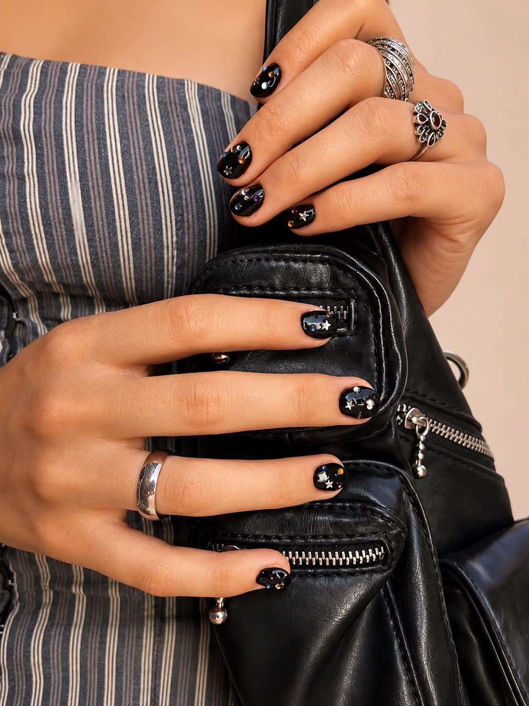 Manos con las uñas pintadas de negro sosteniendo un bolso de cuero negro, con anillos plateados y una blusa a rayas.