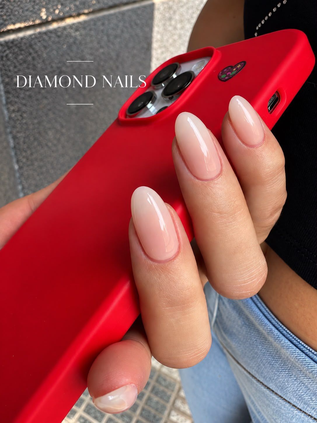 Una mano sostiene un teléfono rojo y luce unas uñas largas, almendradas y de color nude, adornadas con brillantes detalles de diamantes.