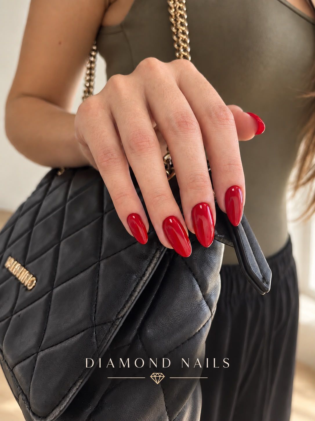 Mano con uñas rojas largas y brillantes apoyada sobre un bolso negro acolchado, con el texto “Diamond Nails”.