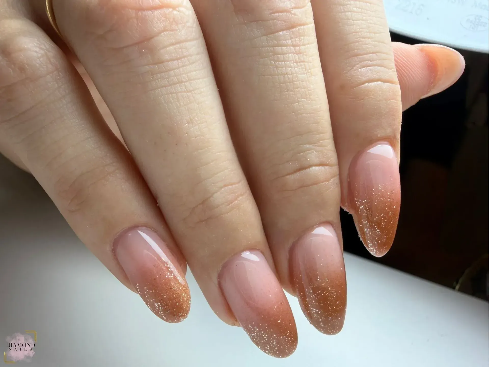 Mano con uñas de forma ovalada que presentan un efecto ombré brillante de color marrón a transparente.