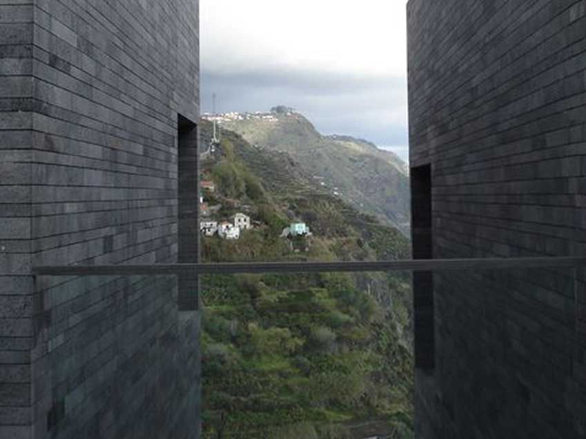 Duas paredes de tijolos escuros emolduram a vista de uma montanha verde com pequenas construções, e um céu nublado