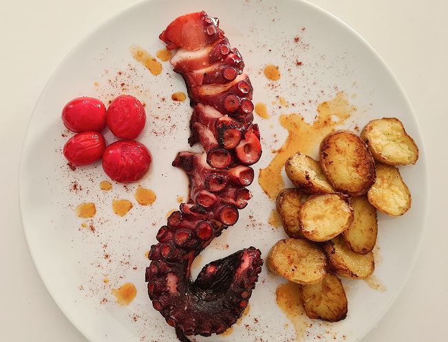 Tentáculo de pulpo a la parrilla con patatas y tomates asados, servido en un plato blanco.