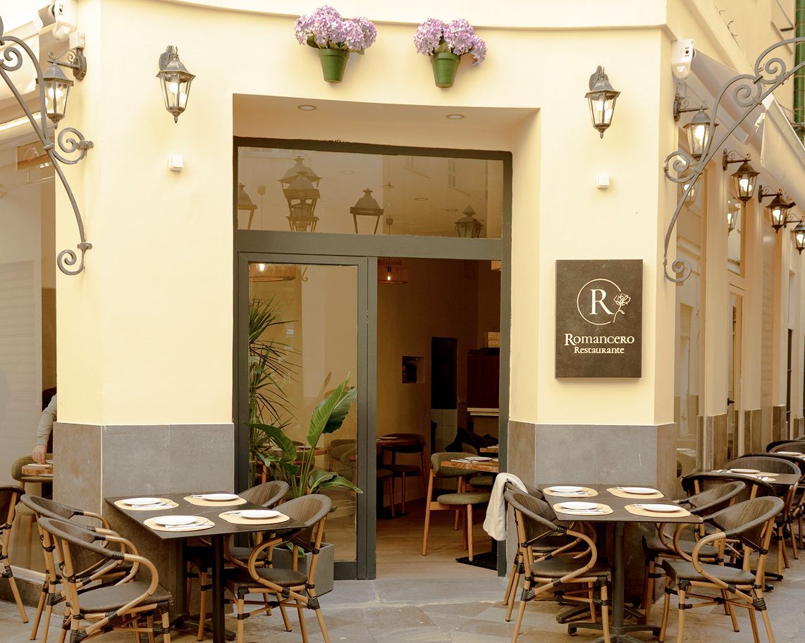 Entrada del restaurante con terraza. Fachada beige, puerta de cristal abierta, mesas oscuras con sillas