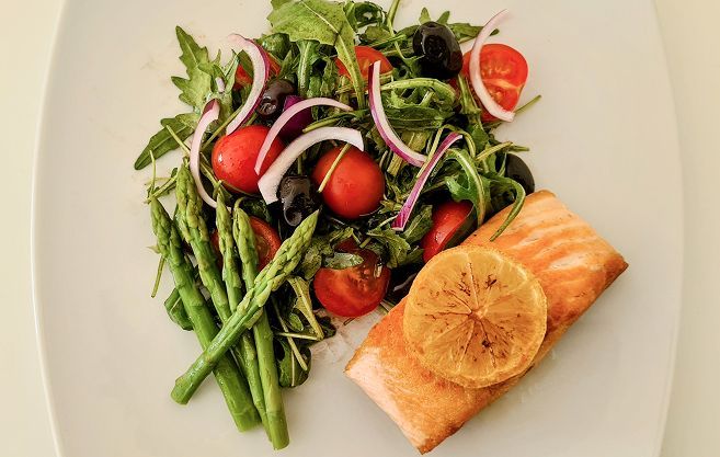 Filete de salmón con rodaja de limón, espárragos y ensalada de rúcula, tomates, cebolla roja y aceitunas en un plato blanco.