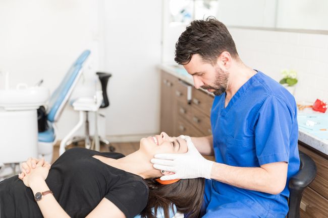 Un dentista examina el rostro de un paciente en su consultorio. El paciente está recostado y sonríe.