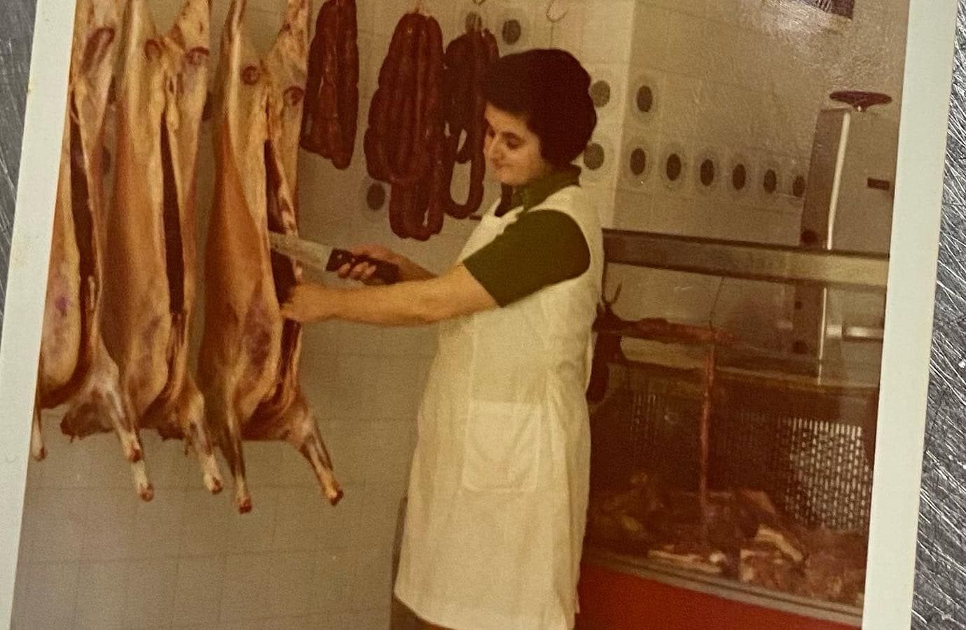 Un carnicero con un delantal blanco corta carne en una tienda, con cadáveres colgando a la izquierda.