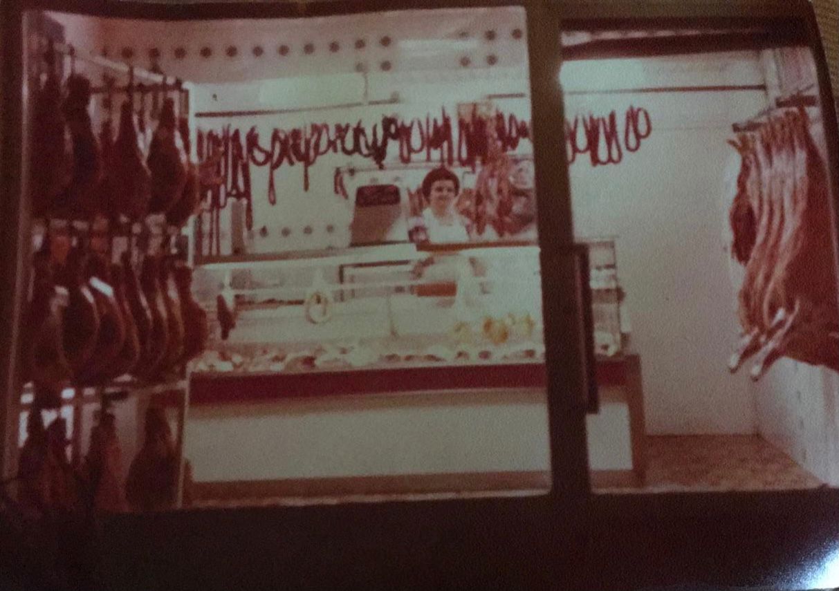 Una mujer se encuentra dentro de una carnicería, detrás de una vitrina llena de carne, con filas de embutidos colgando.