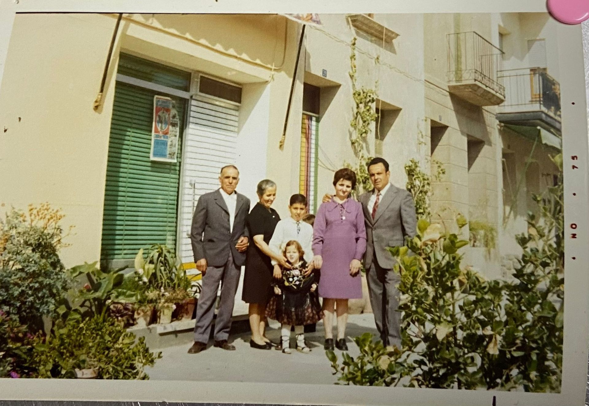 Familia posa afuera de un edificio con persianas verdes, probablemente una casa, con vegetación, Italia.