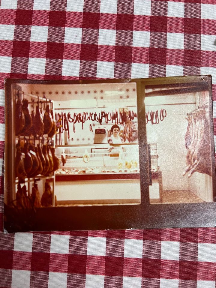 Interior de carnicería con carnes colgadas, una persona detrás del mostrador y un mantel a cuadros rojos y blancos.