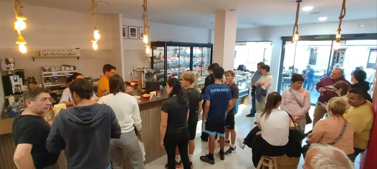 Fotografía interior de una cafetería con gente esperando. Mostrador, luces y algo de comida visibles.