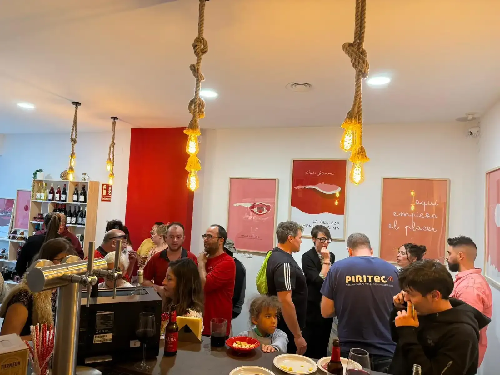 Gente en un bar, algunos bebiendo, otros charlando. Interior con paredes rojas y blancas y luces colgantes.