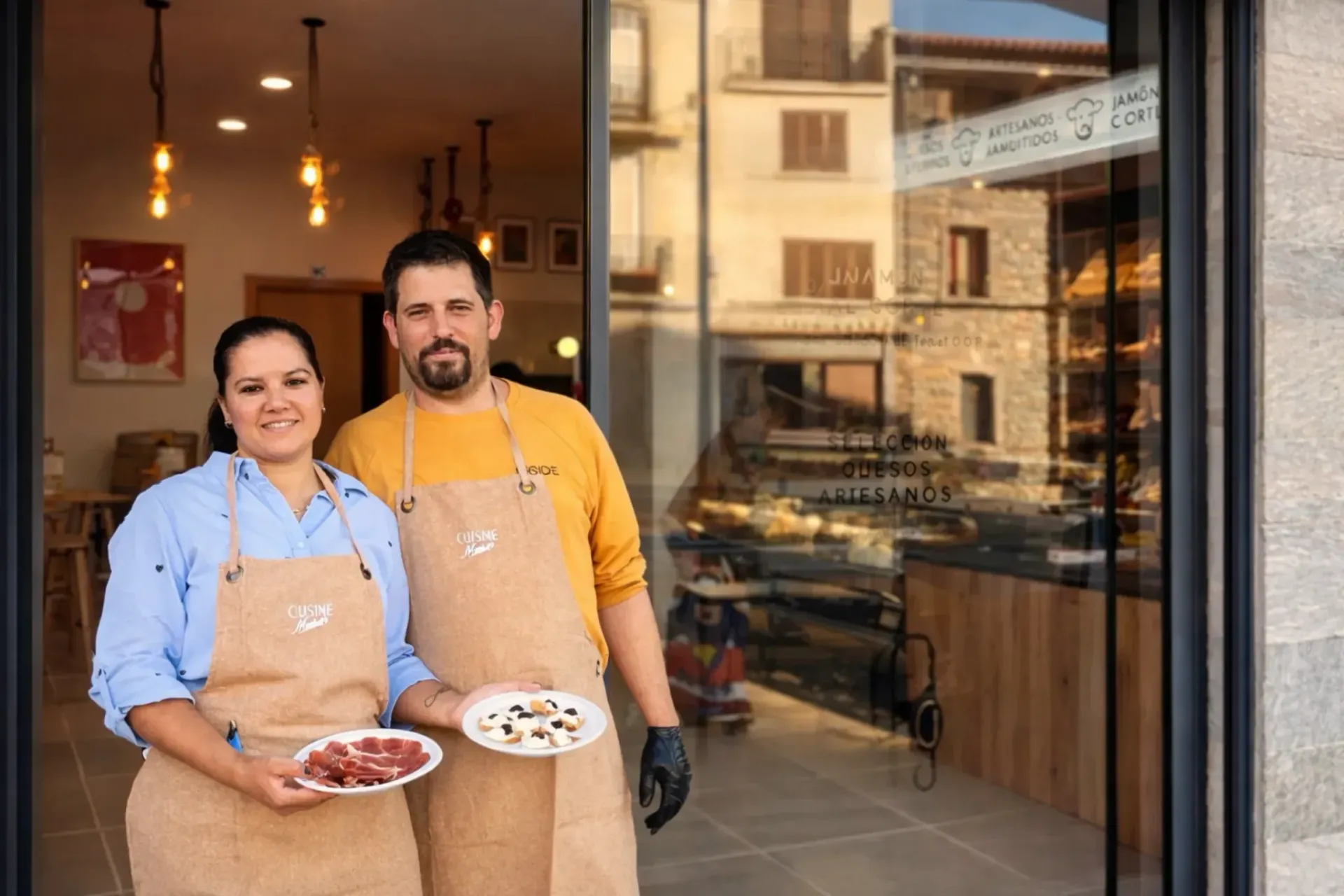 Dos personas con delantales están paradas en la puerta de una tienda, cada una sosteniendo platos de comida.