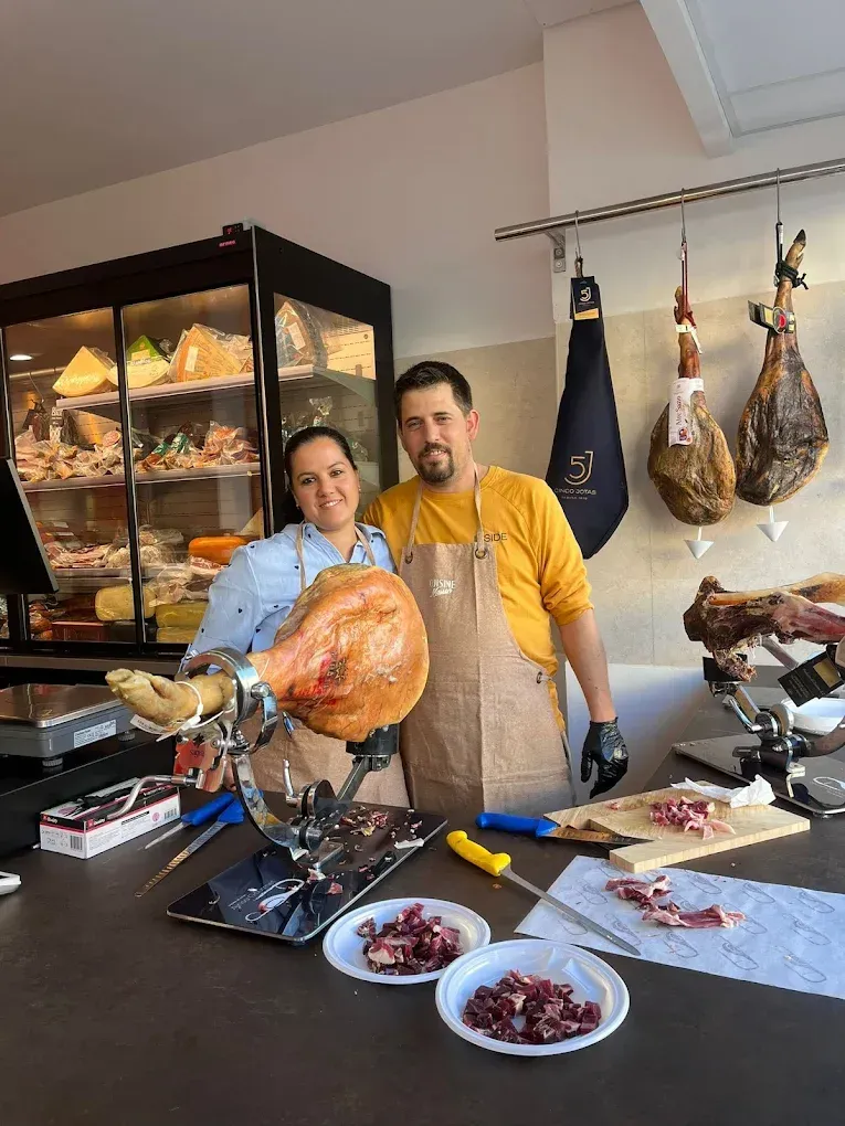 Dos personas posan tras un mostrador con un jamón curado. Se ve la exposición de embutidos y quesos.