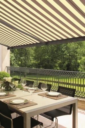 Esstisch im Freien mit gestreifter Markise, Blick auf ein schmiedeeisernes Geländer und Grünanlagen.