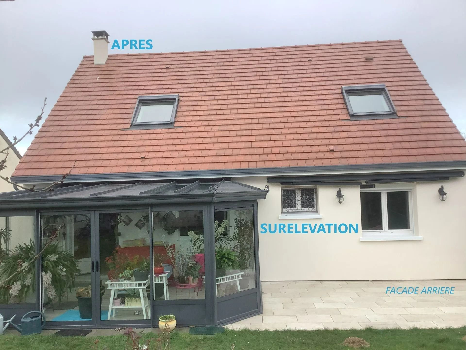 Une maison avec un toit de tuiles rouges, deux puits de lumière, une véranda et un bardage beige.