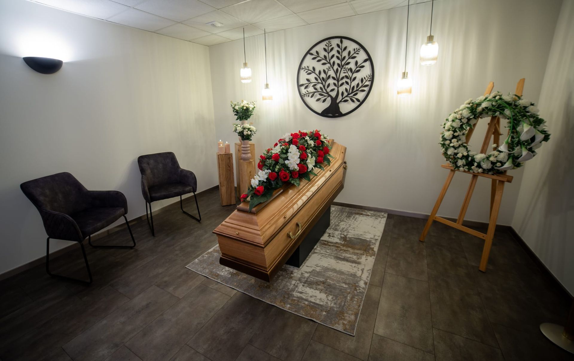 Un cercueil en bois clair orné de fleurs rouges et blanches, placé dans une pièce calme avec des chaises et une décoration murale circulaire.