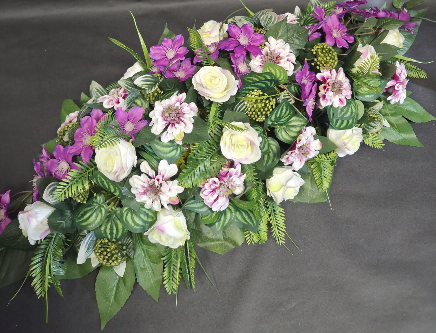 Une composition florale allongée mettant en scène des roses blanches, des clématites violettes et du feuillage vert sur un fond gris foncé.