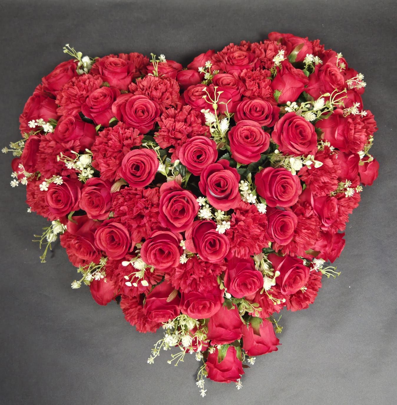 Une composition florale en forme de cœur, réalisée avec des roses rouge foncé, des œillets et de petites fleurs blanches d'accompagnement, sur un fond gris.