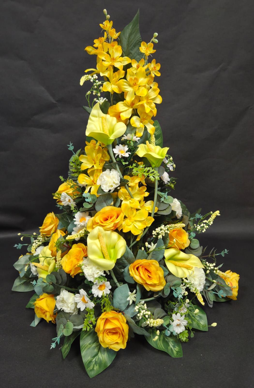 Une composition florale triangulaire et élancée, composée de roses jaunes, de callas et de fleurs blanches en accentuation, sur fond noir.