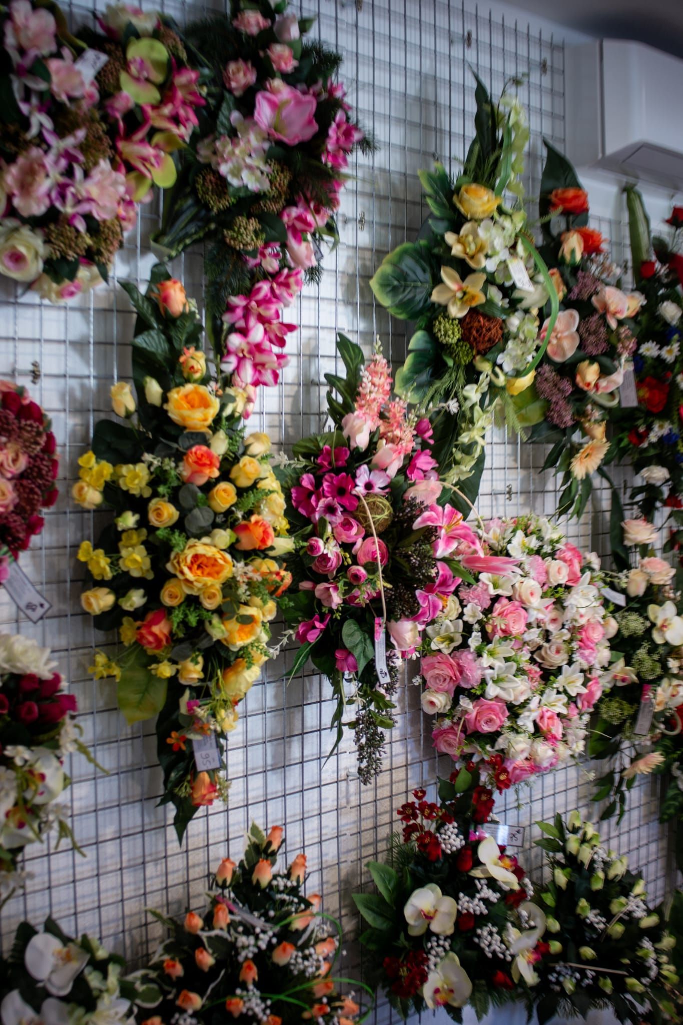 Un étalage de diverses compositions florales artificielles colorées est suspendu à un mur à grille blanche.