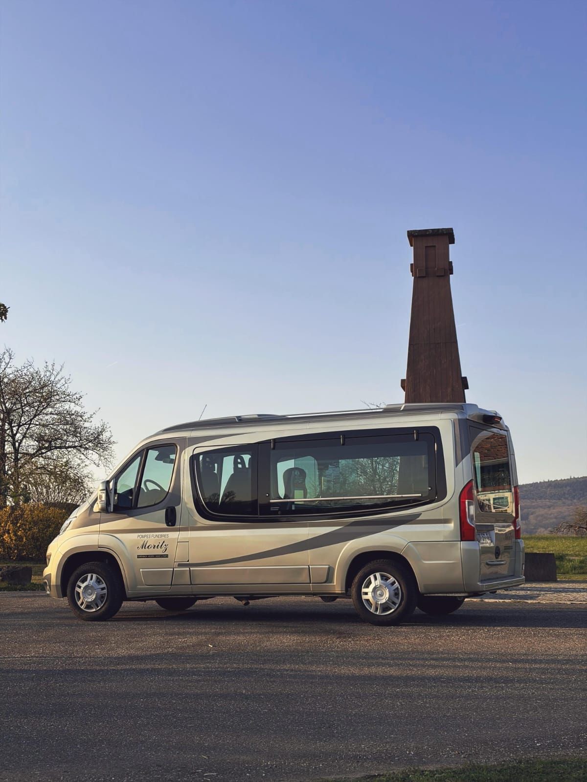 Un camping-car argenté garé sur du gravier devant une vieille tour en pierre, sous un ciel bleu azur.