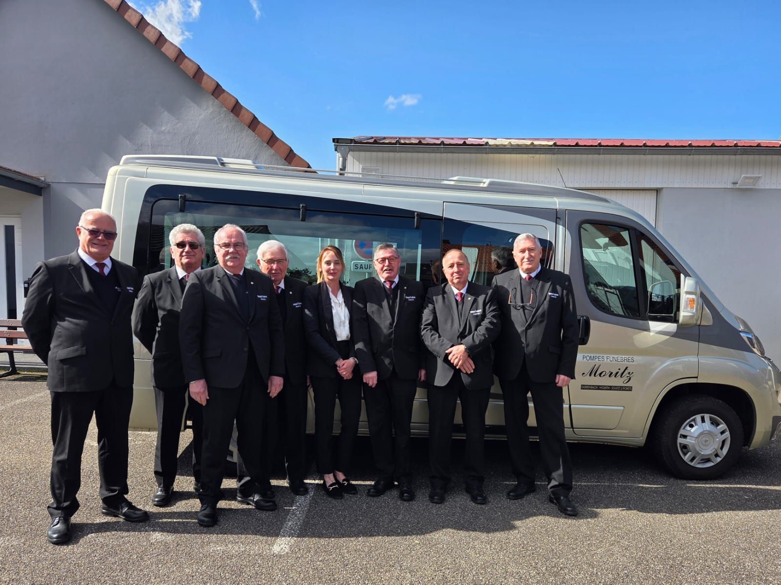Un groupe de huit professionnels en uniformes sombres assortis, alignés devant un corbillard clair à l'extérieur.