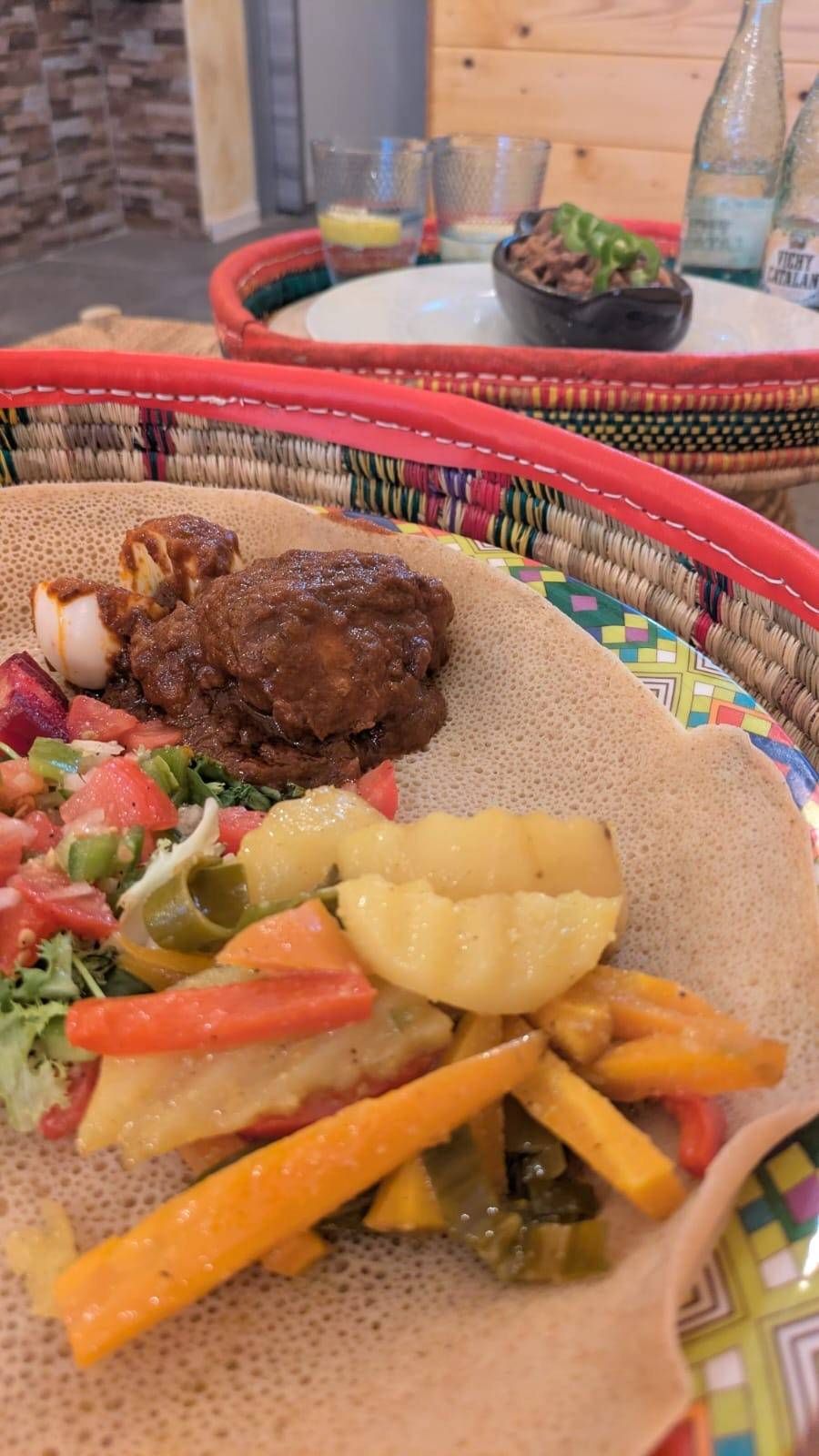 Comida etíope en pan injera, servida en una cesta tejida. Incluye carne, verduras y ensalada.