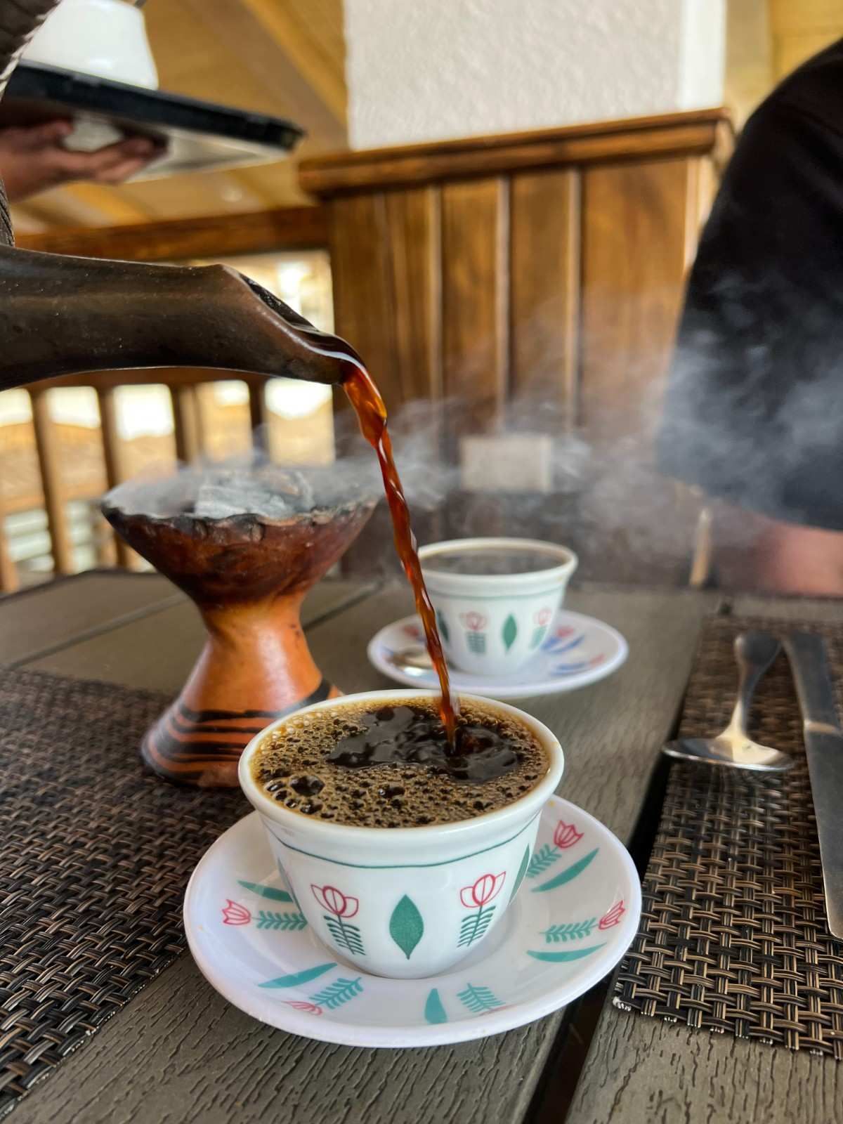 Se vierte café en una taza sobre una mesa, mientras sale humo de un quemador de incienso cercano.