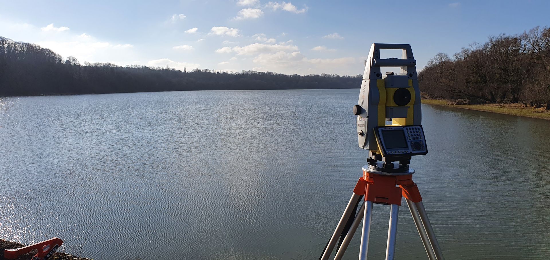 Scanner topographique sur trépied au bord d'un lac calme sous un ciel partiellement nuageux.