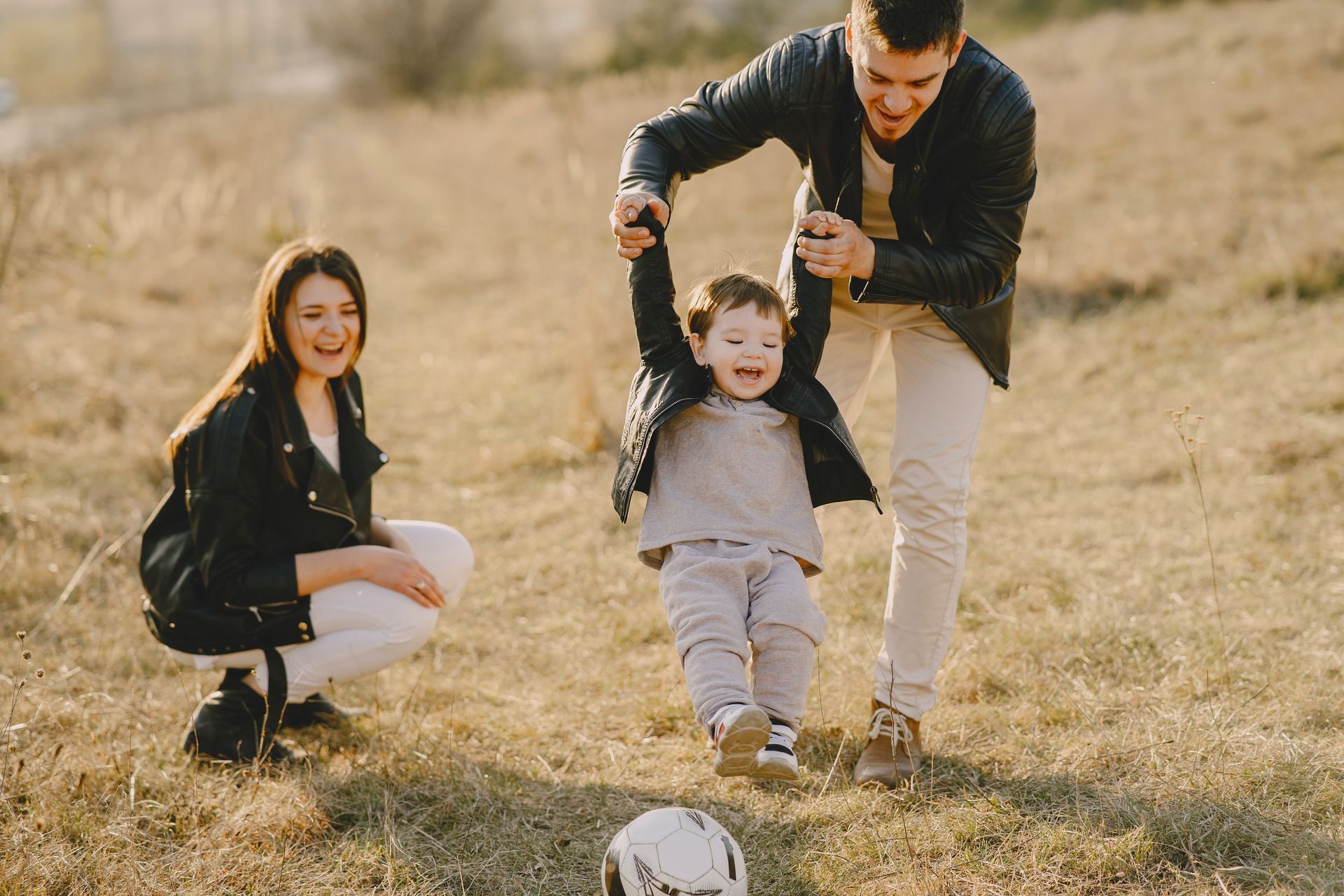 Familia jugando al aire libre: Padre balanceando a un niño, madre arrodillada cerca y sonriendo en un campo.