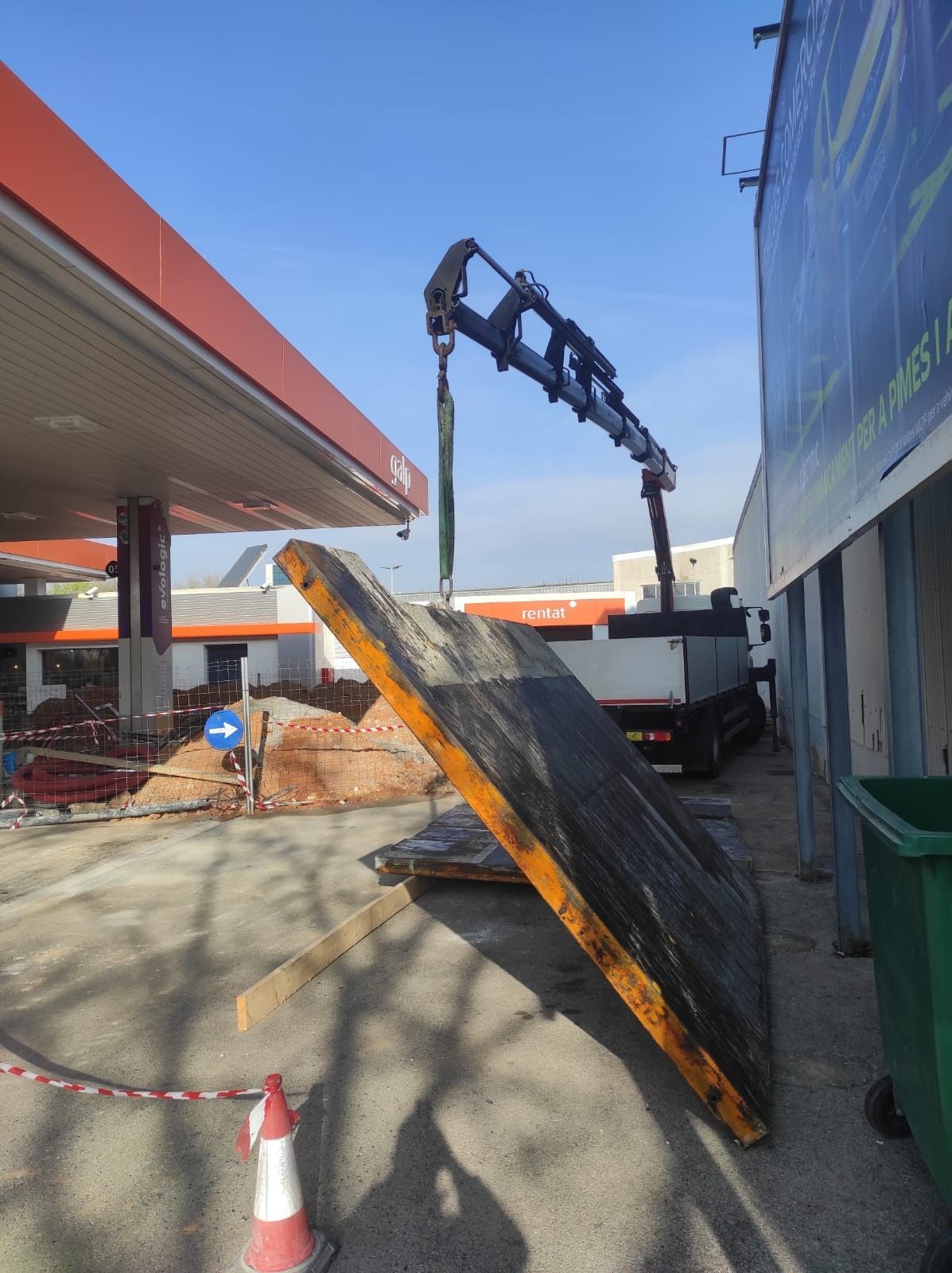 Una grúa está levantando un gran trozo de hormigón frente a una gasolinera.