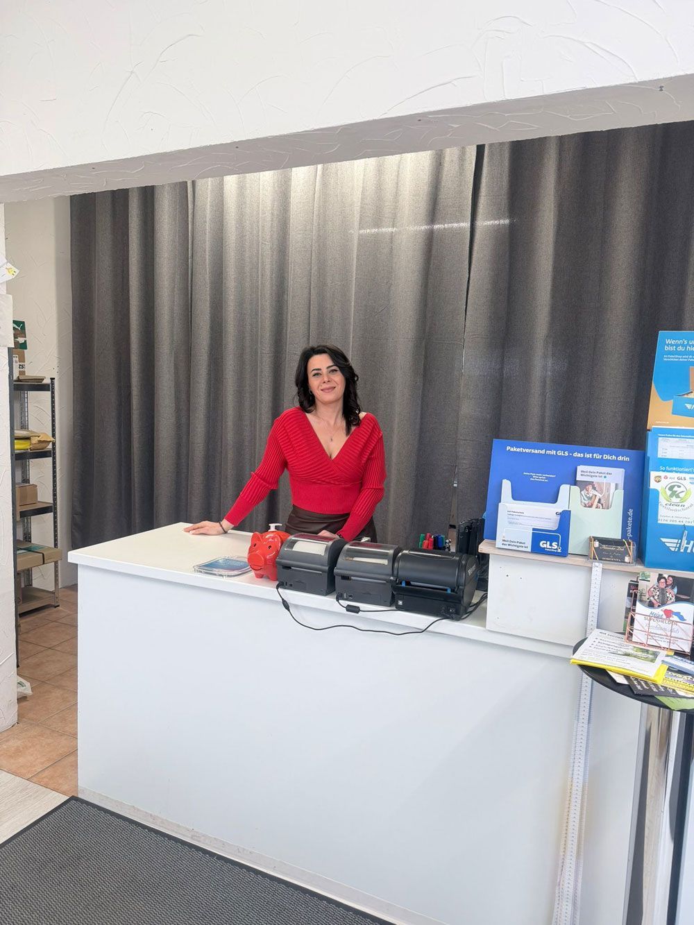 Eine Frau in einem roten Oberteil steht hinter einem weißen Tresen mit Geräten; im Hintergrund sind graue Vorhänge zu sehen.