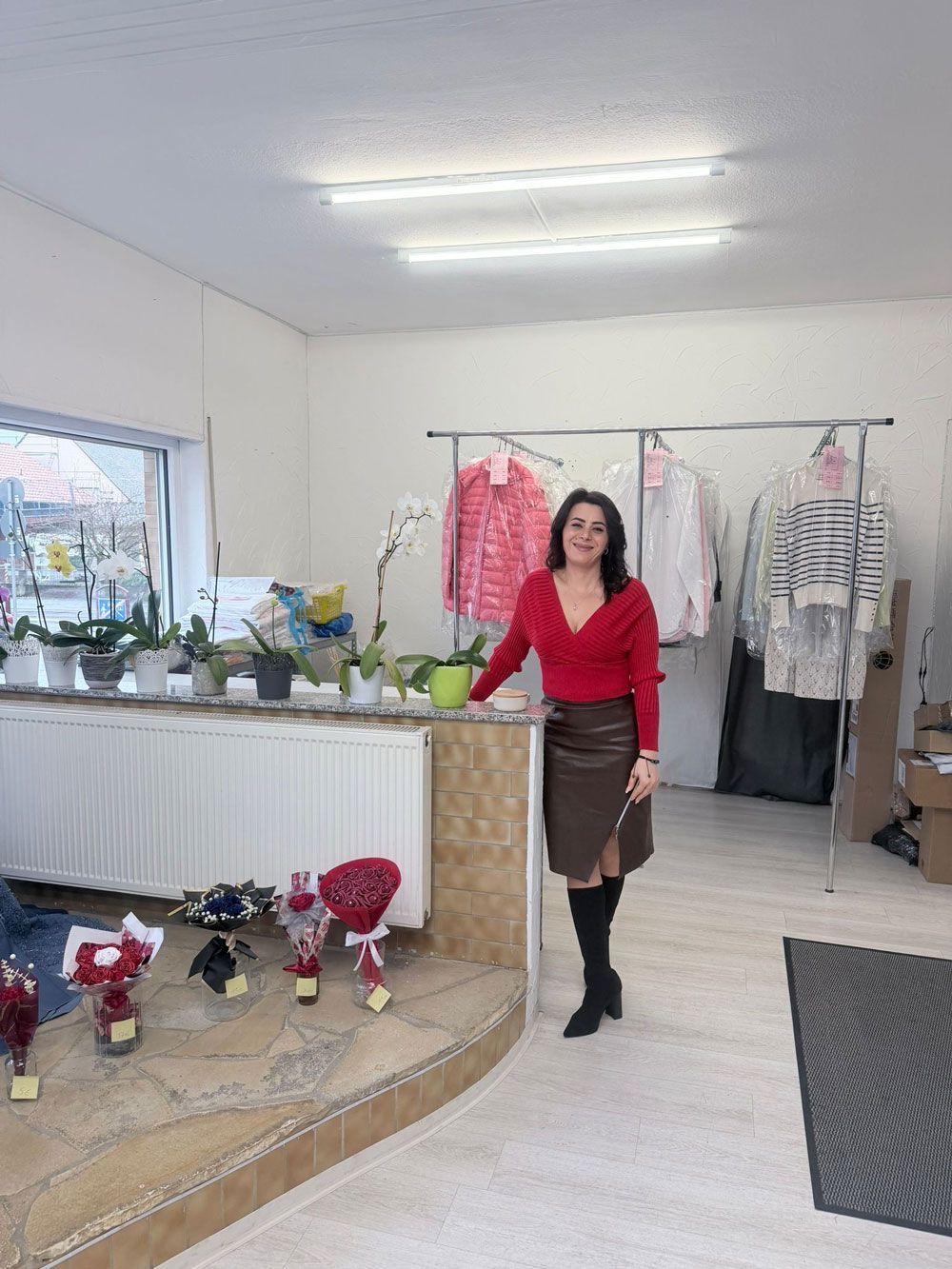 Eine Frau in rotem Oberteil und braunem Rock steht mit Blumen in einem Bekleidungsgeschäft.