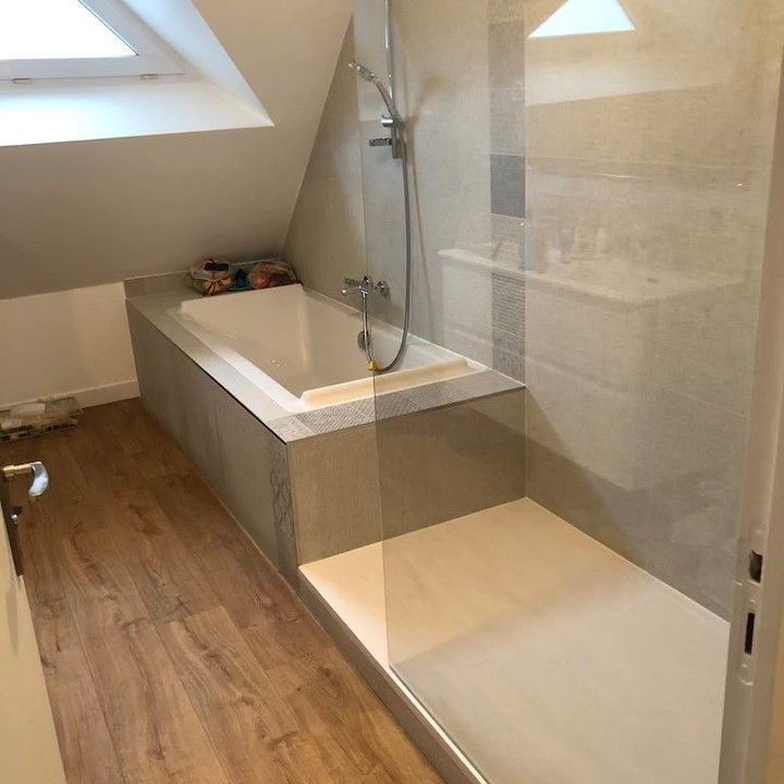 Salle de bains mansardée avec baignoire et douche. Sol imitation bois et murs carrelés gris.