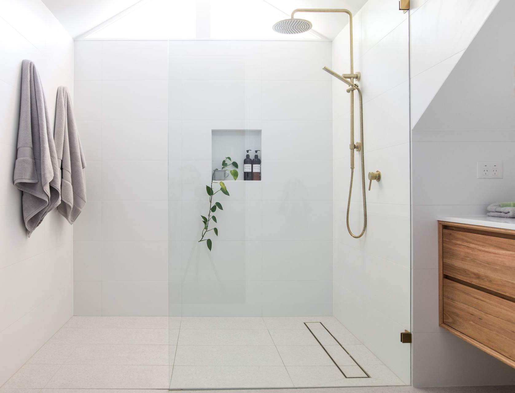 Douche moderne blanche avec robinetterie dorée, paroi de verre, sol carrelé et meuble en bois.