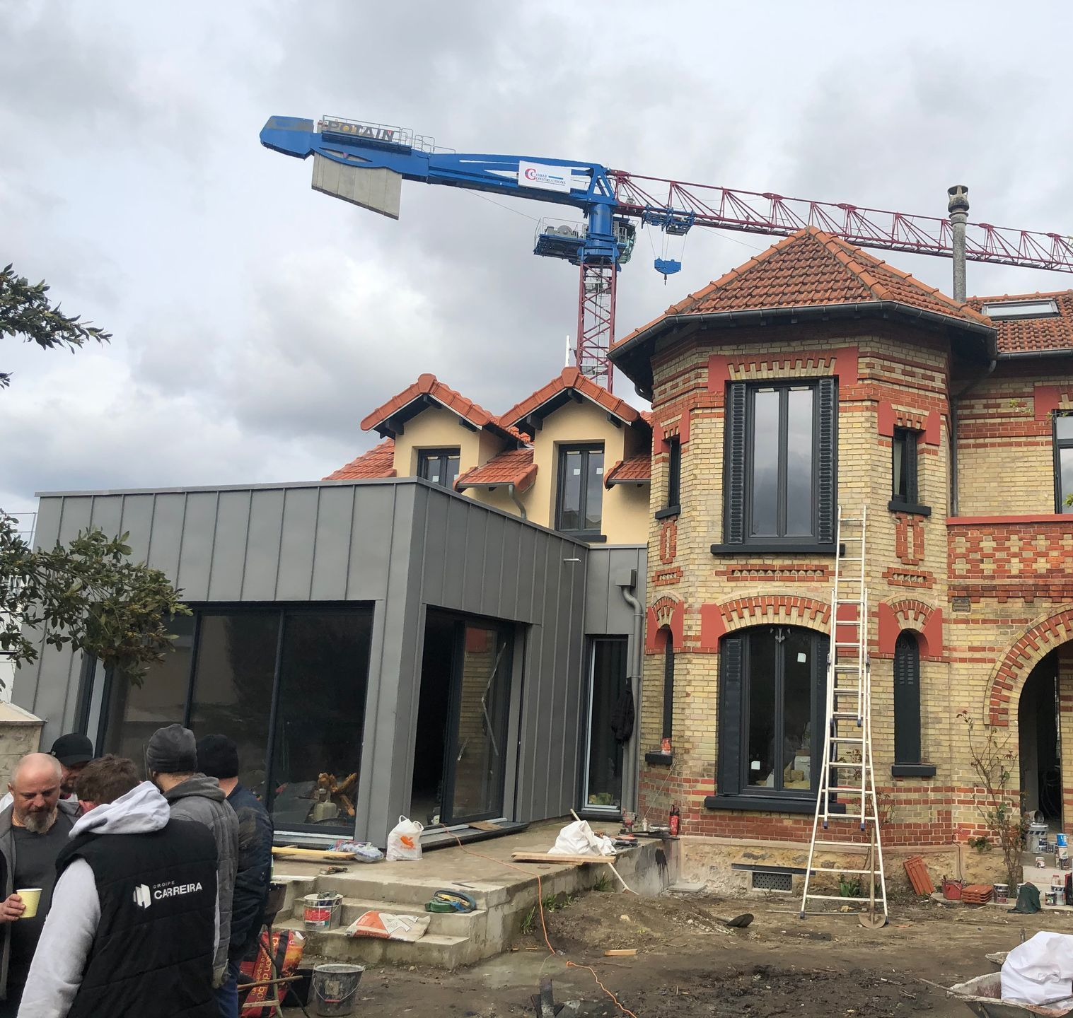 Chantier de construction avec un bâtiment en briques de deux étages, une extension grise et une grue ; plusieurs personnes se tiennent à proximité.