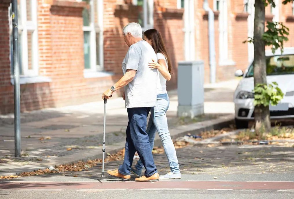 Una mujer está ayudando a un anciano con un bastón a cruzar la calle.