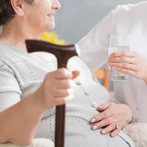 Una mujer mayor sostiene un bastón y un vaso de agua.