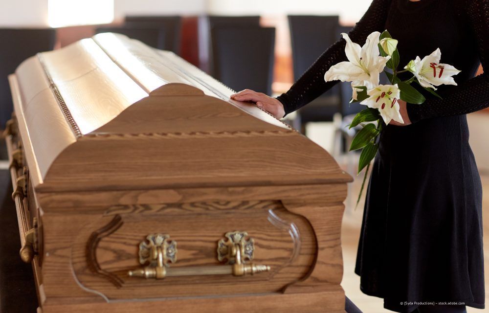 Ein Holzsarg bei einer Beerdigung, daneben eine Person in Schwarz, die weiße Lilien hält.