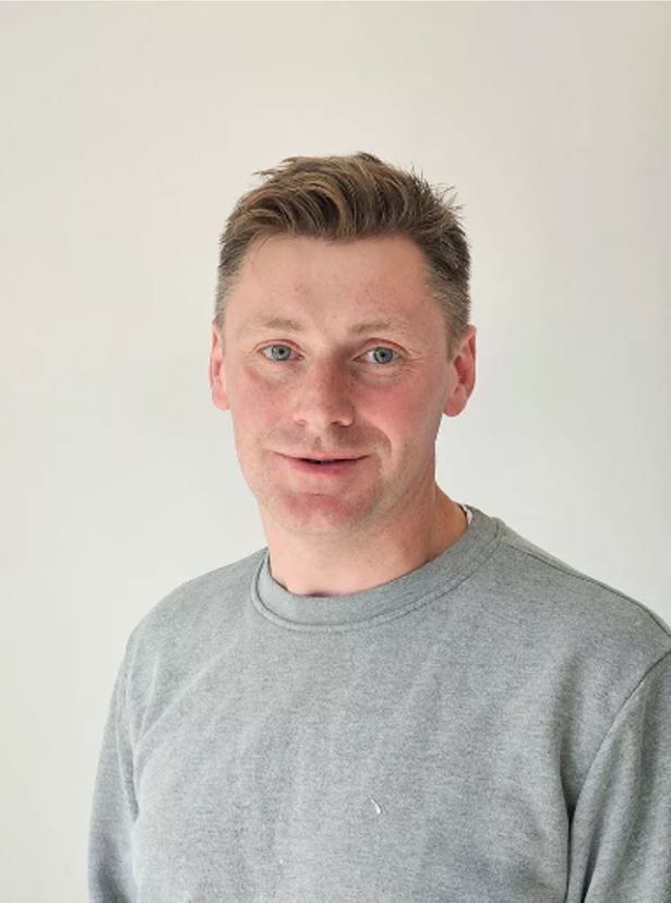 Ein Mann in einem grauen Pullover lächelt in die Kamera.