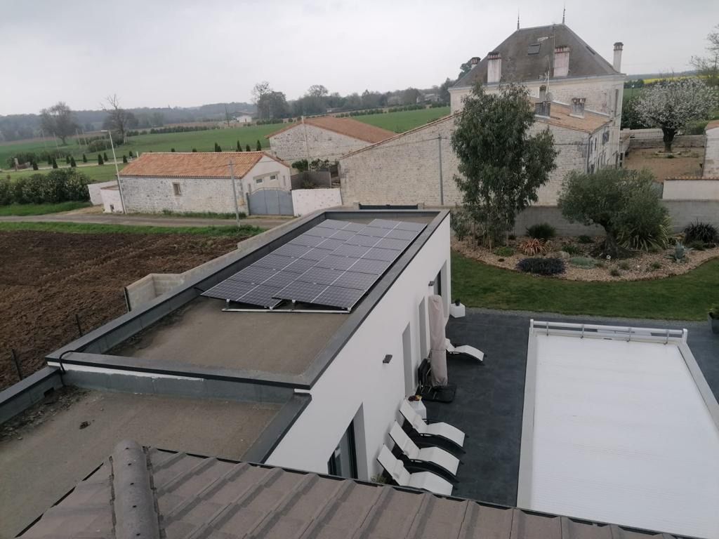 Panneaux solaires sur le toit plat d'un bâtiment blanc, en milieu rural. Autres bâtiments et champs à l'arrière-plan.