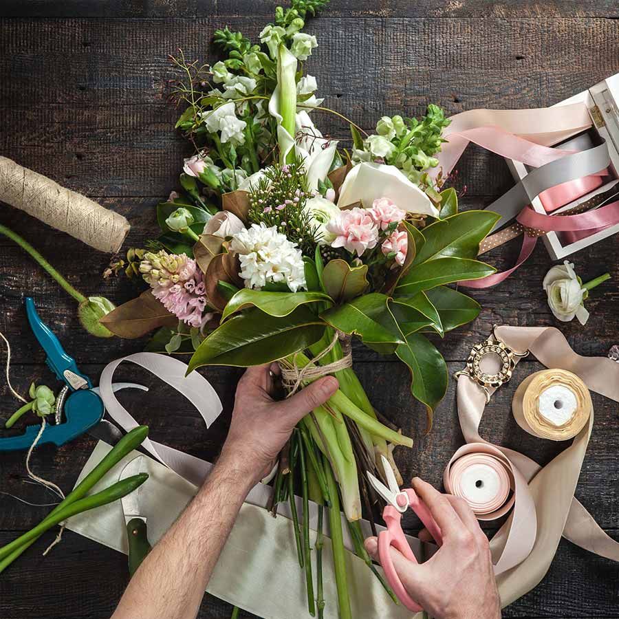 Unas manos arreglan un colorido ramo de flores sobre una mesa de trabajo con cintas, tijeras y velas.