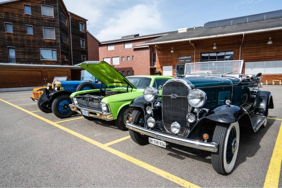 Drei Oldtimer stehen auf einem Parkplatz; ein grünes, ein schwarzes und ein blaues Fahrzeug. Im Hintergrund sind Gebäude zu sehen.