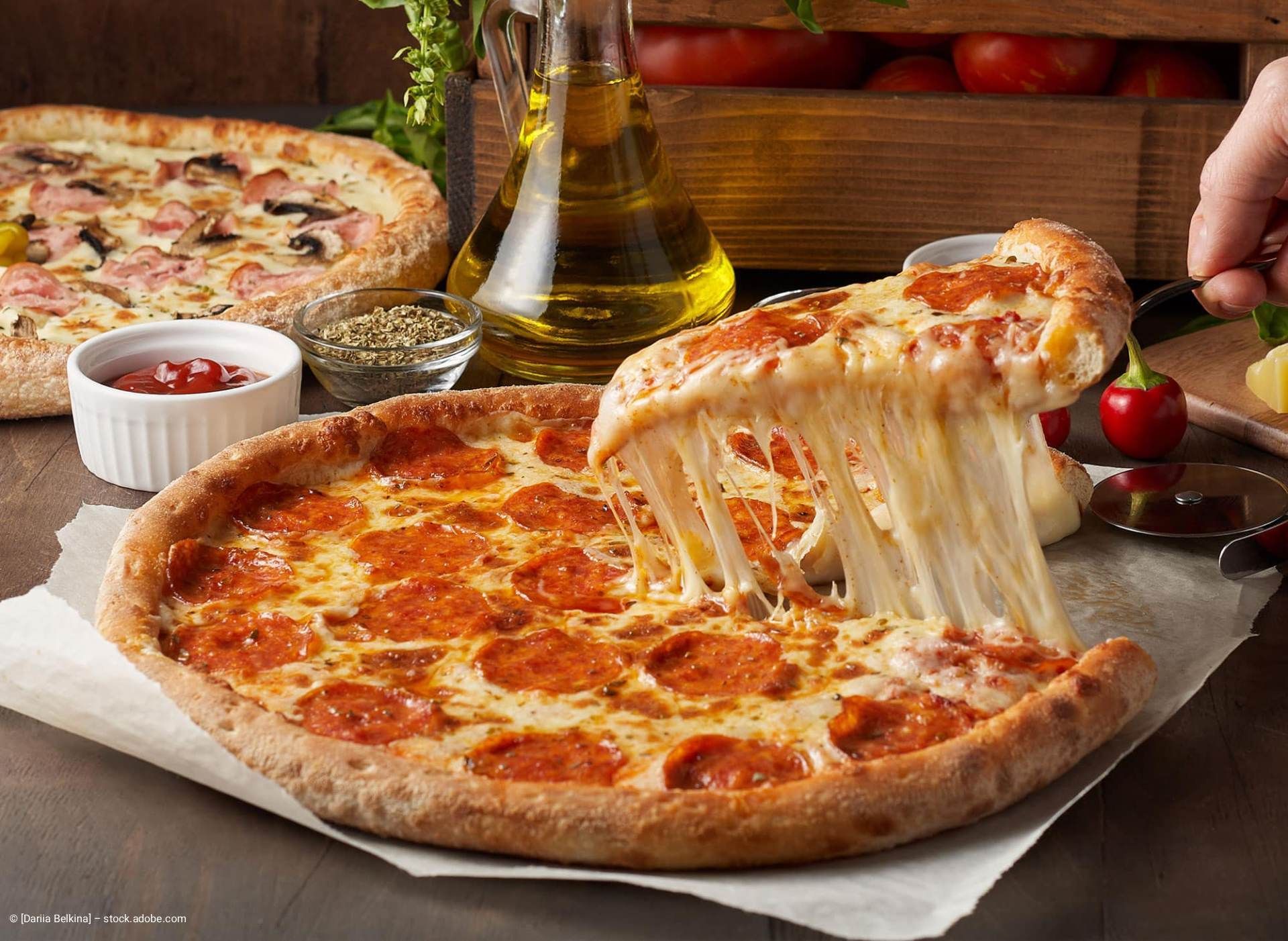 Ein Stück Pepperoni-Pizza mit gezogenem Käse, weitere Pizza, Öl, Kräuter und Soße stehen auf dem Tisch.